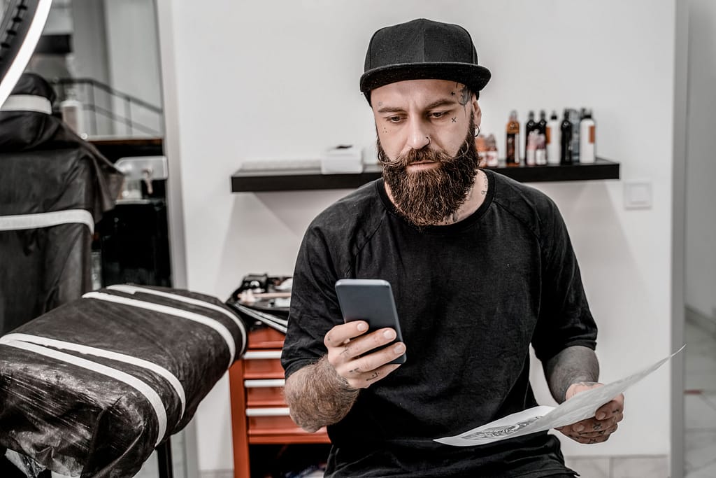 young-male-tattoo-artist-with-beard-holding-phone-sketch-sitting-couch-workshop-place